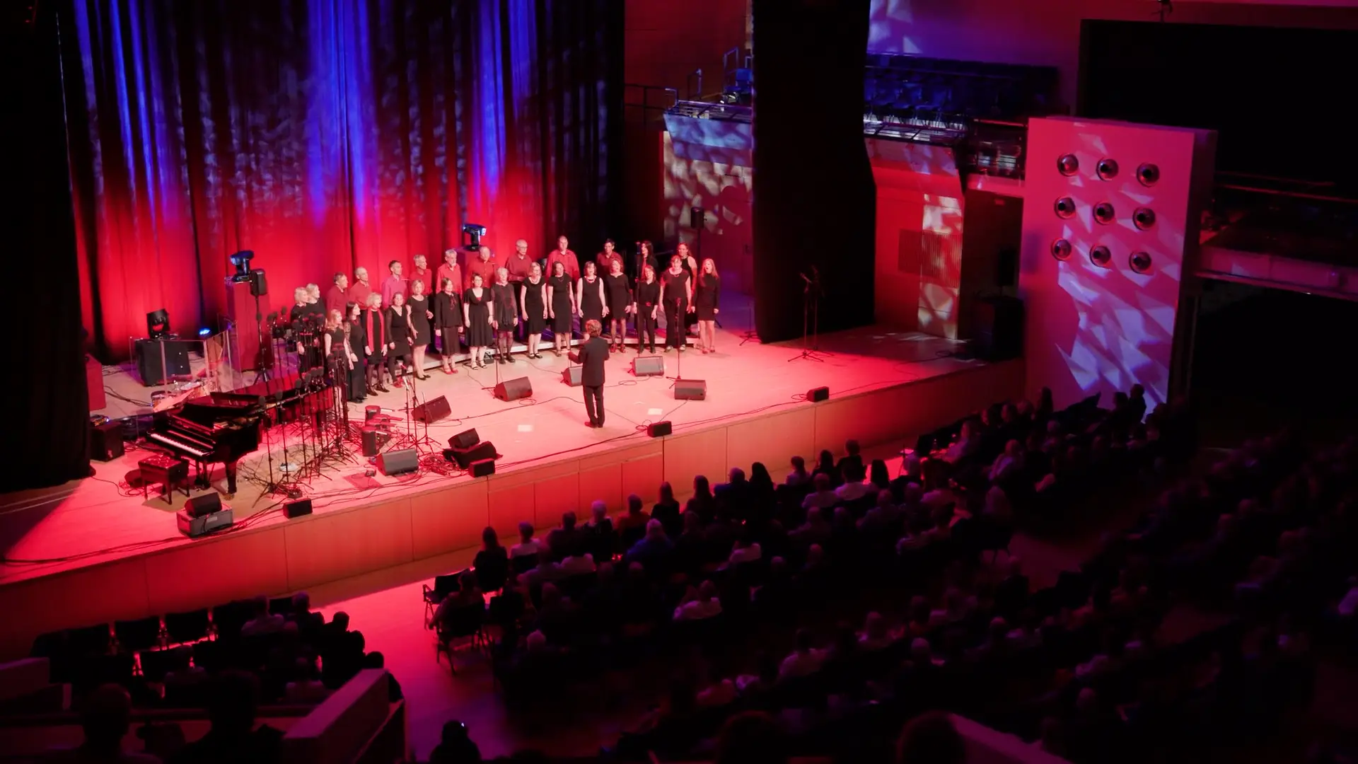 Ein Chor steht auf einer Bühne. Ein Dirigent steht mit dem Rücken zu einem Publikum. Das Publikum ist voller, mit nur wenigen freien Sitzen in der Händelhalle.