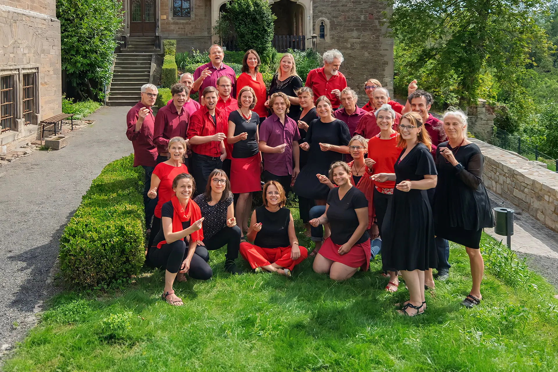 Jazzchor aus Halle steht gemeinsam im Garten vor historischem Gebäude, Mitglieder in roten und schwarzen Outfits